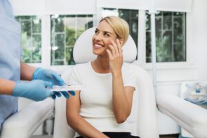 blonde woman sits in chair touching face and smilling with doctor standing next to her